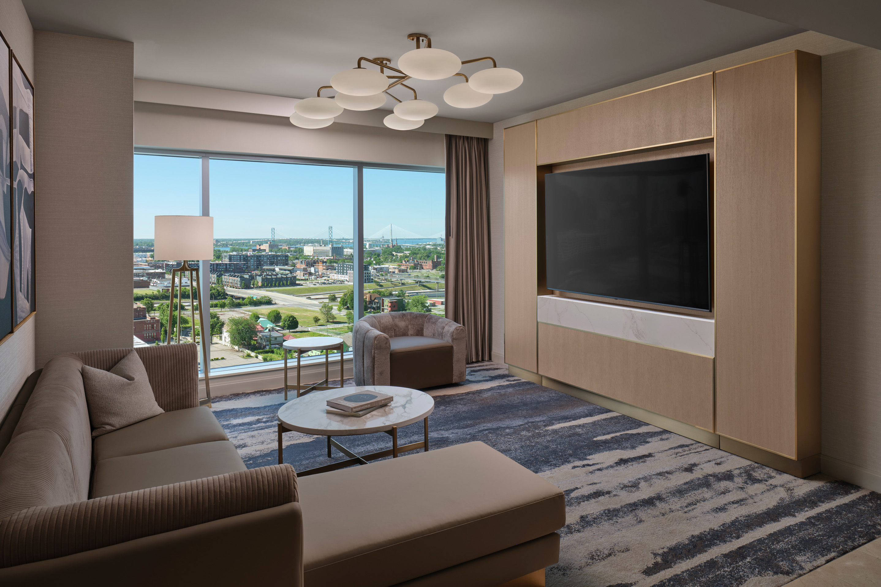 Luxury hotel suite living room with sofa, large TV, and floor-to-ceiling windows overlooking Detroit