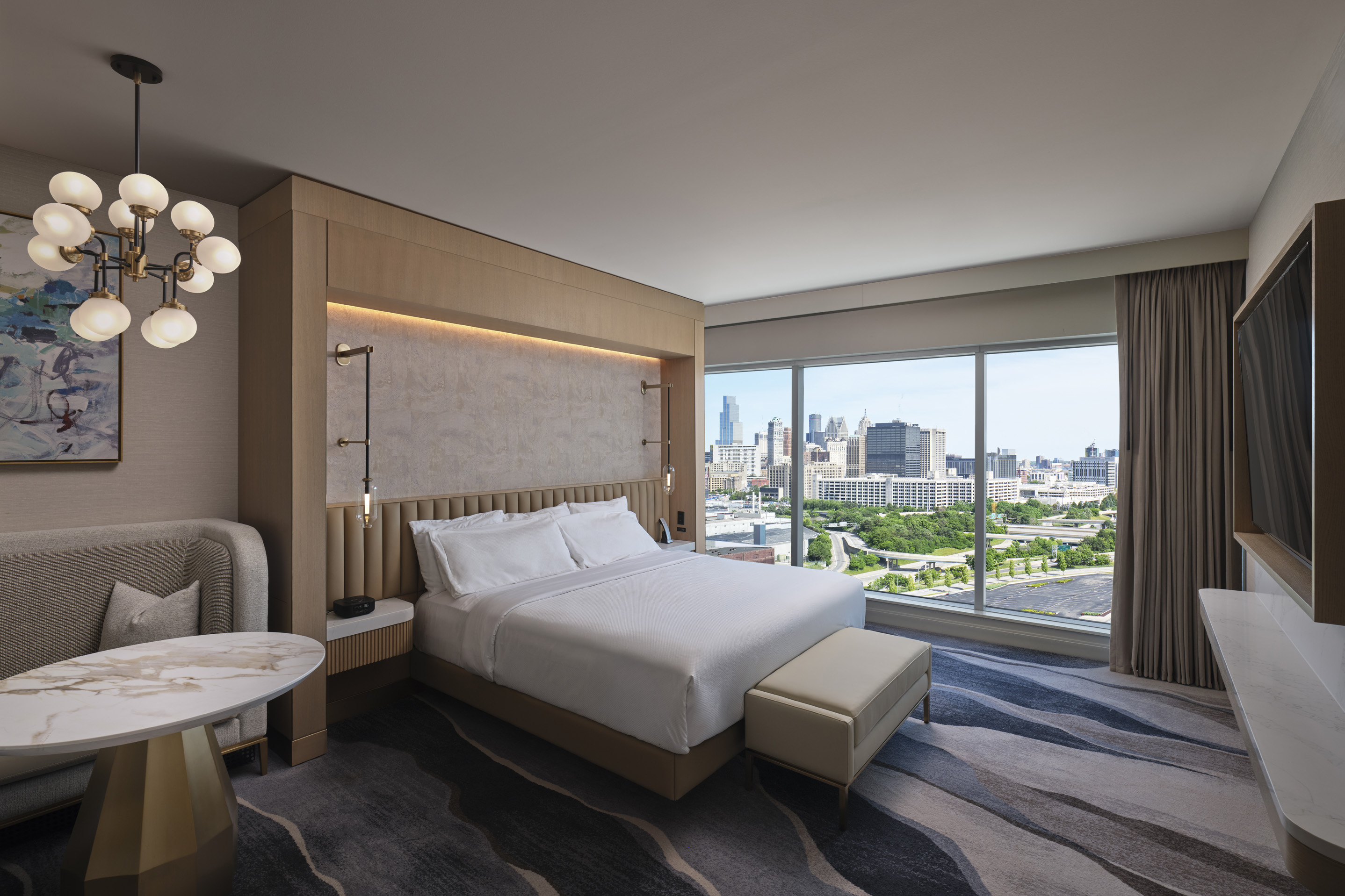 Luxury hotel room with king bed, modern lighting, and floor-to-ceiling windows overlooking Detroit